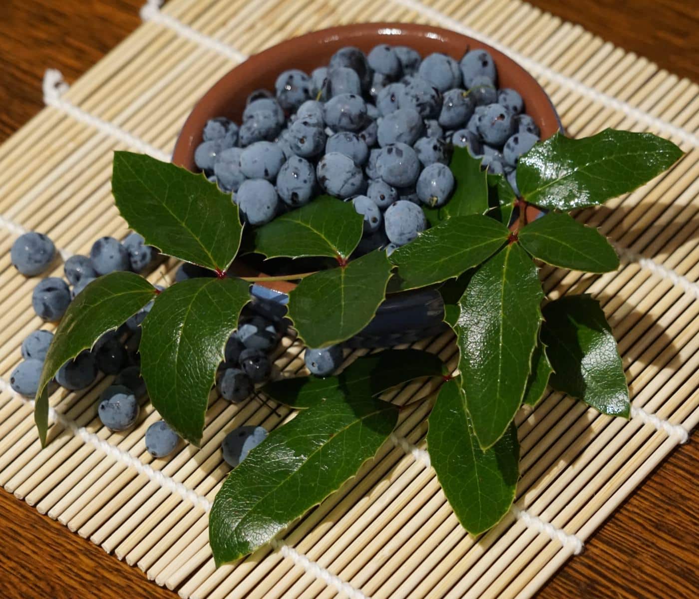 Bowl of Mahonia berries with leaves on wicker mat.