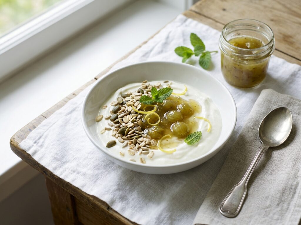 A white bowl of thick yogurt topped with glossy green-gold gooseberry compote, scattered toasted seeds and oats, lemon zest and fresh mint leaves